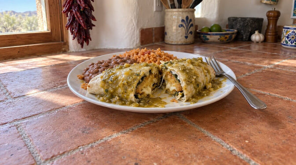 A flour tortilla burrito cut in half and smothered in green chile sauce and melted cheese on a white plate, with refried beans and rice, on a tile kitchen counter