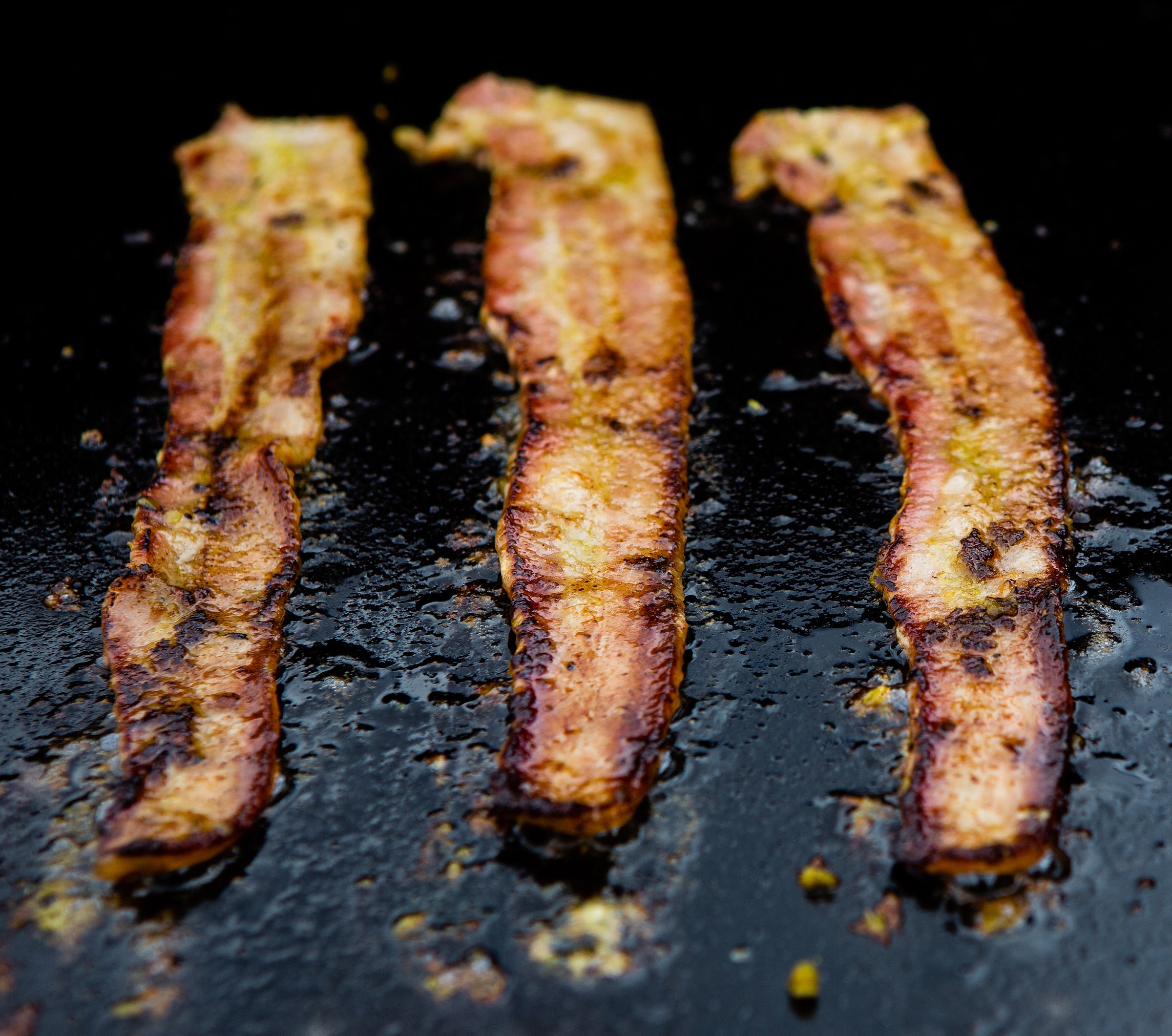 Three strips of crispy Applewood Smoked Bacon frying on a black skillet, with glistening fat and golden-brown textures.