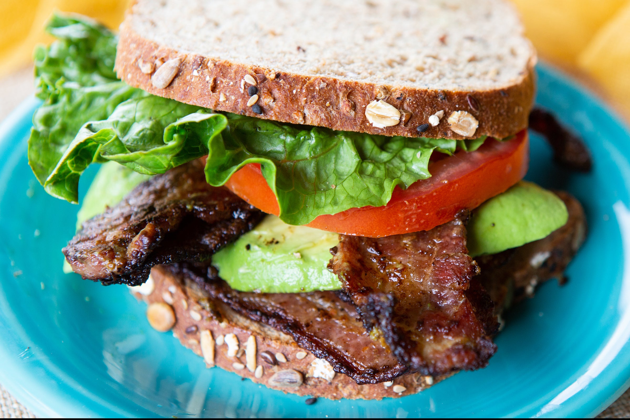 Close-up of a sandwich with whole grain bread, lettuce, tomato, avocado, Maple Leaf Farms Hatch Green Chile Bacon, and grilled meat on a blue plate.