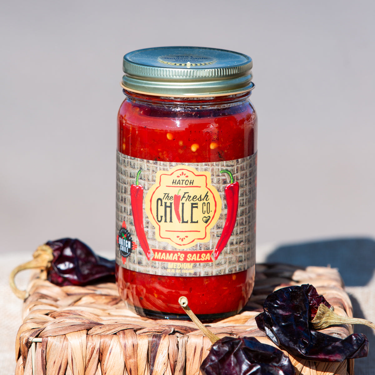 A jar of Fresh Chile Co's Mama's Blended Hatch Chile Salsa sits on a woven mat, surrounded by dried red chili peppers, with a light beige backdrop.