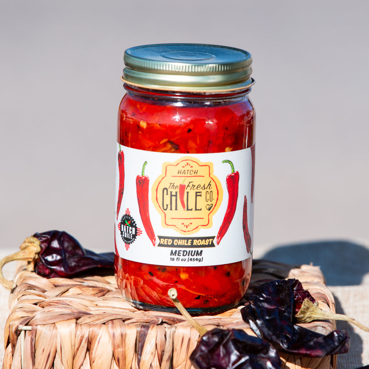 A jar of medium roast Hatch Red Chile Roast from the hatch chile co. on a wicker basket, flanked by dried chili peppers, against a neutral background.