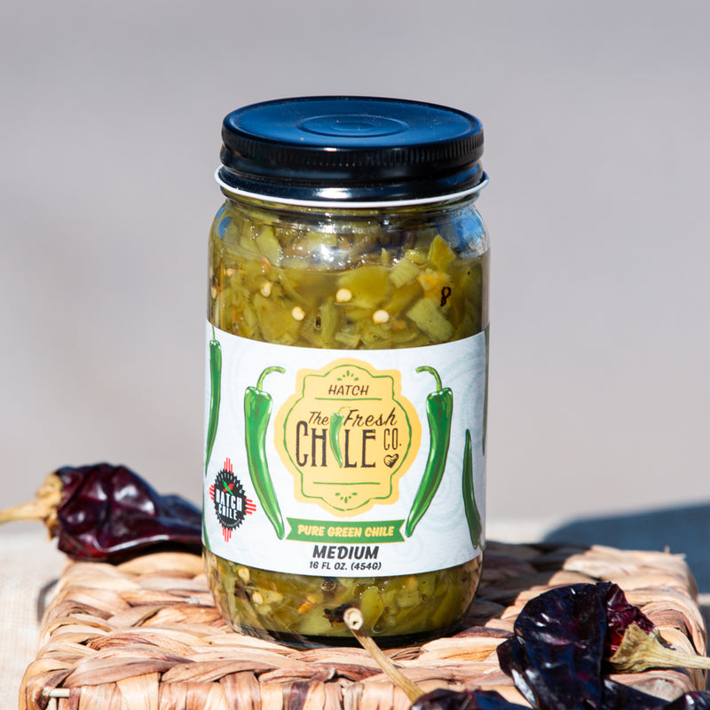 A jar of Pure Hatch Green Chile, labeled as medium heat, placed on a wicker surface with dried chilies scattered around it.