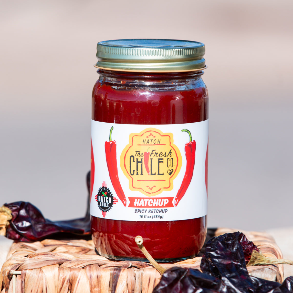 A jar of Spicy Hatch Red Chile Ketchup from The Fresh Chile Co. is displayed, with a label featuring red and green chile peppers. The jar sits on a woven mat accompanied by dried chiles.