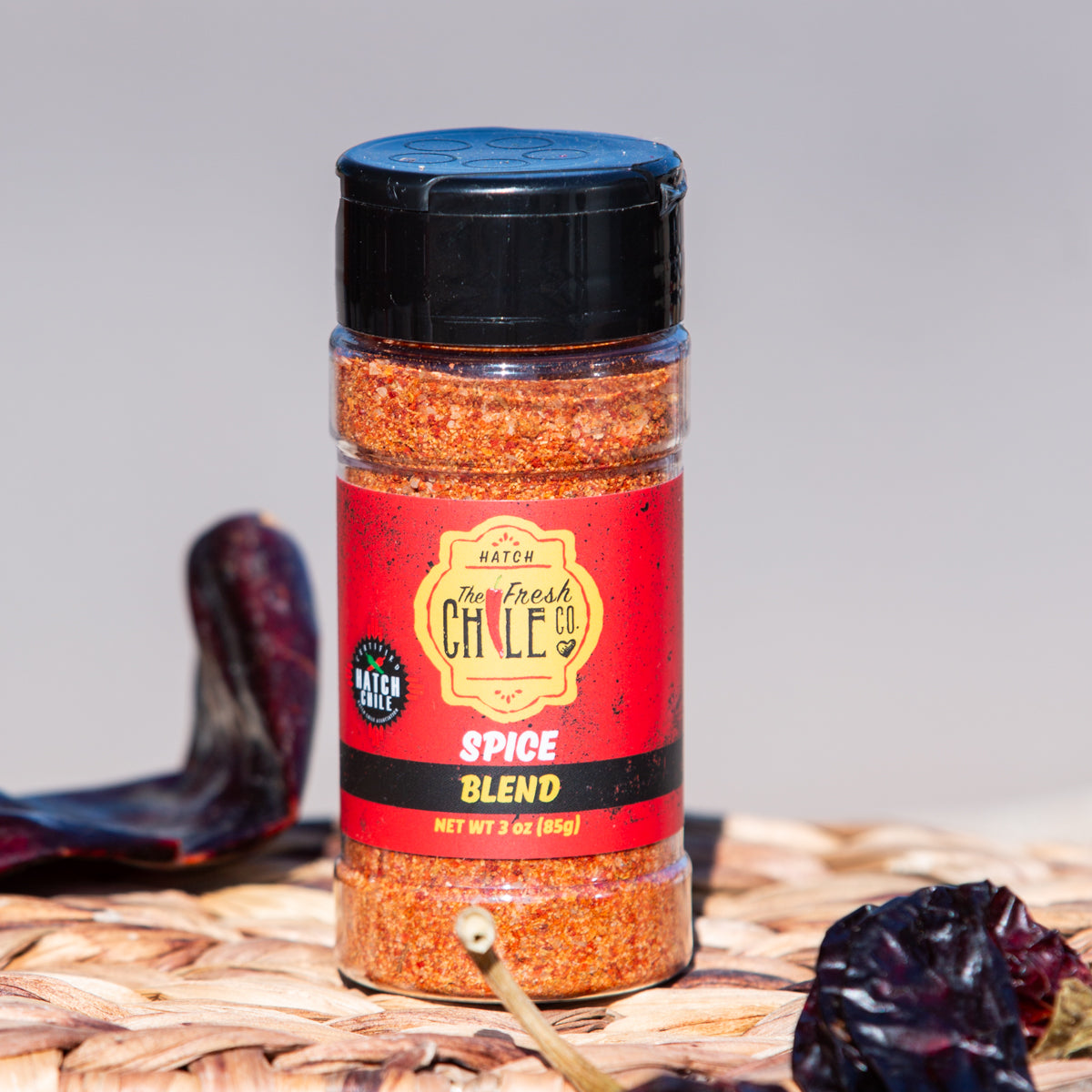 A jar of "the fresh chile co." Hatch Red Chile Spice Blend on a woven mat, with dried red chiles scattered around it. The jar label is vibrant with red and gold colors.