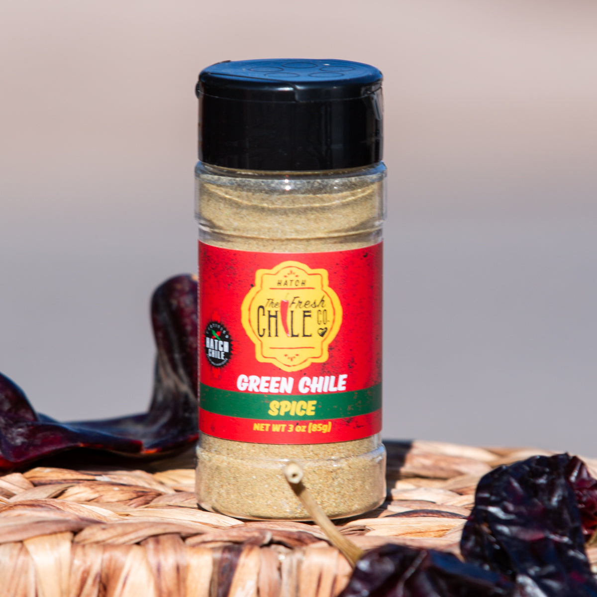 A bottle of Hatch Green Chile Spice placed on a wicker surface, flanked by dried red chilies. The label on the bottle displays vibrant red and yellow colors.