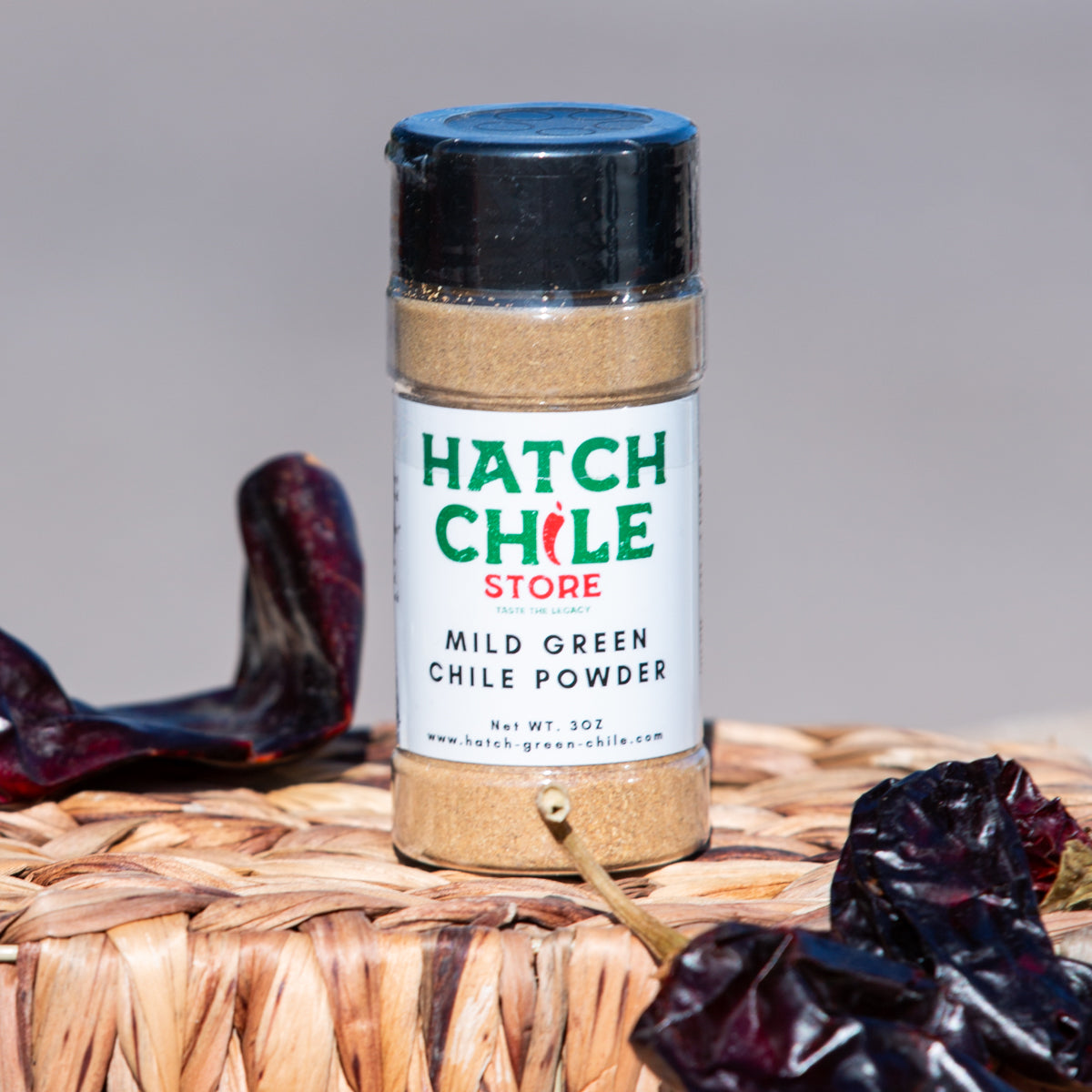 A bottle of Green Chile Powder, surrounded by dried red chiles on a woven mat, against a neutral background.