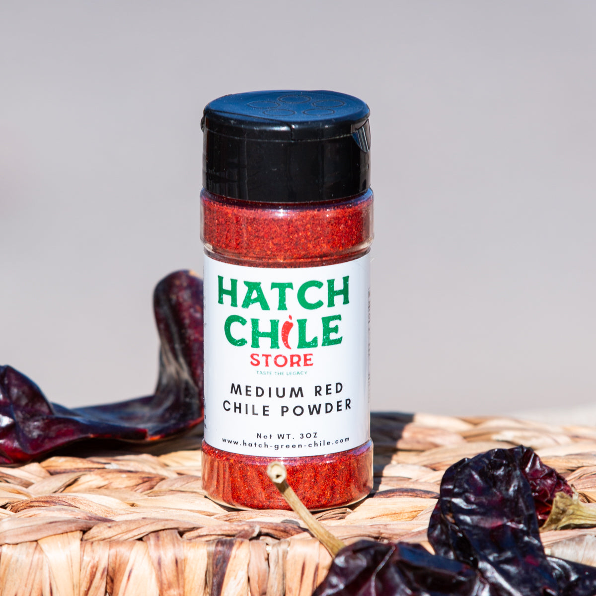 A container of Red Chile Powder, positioned in the center, surrounded by sun-dried red chilies on a woven surface. The background is plain, enhancing the vivid colors.