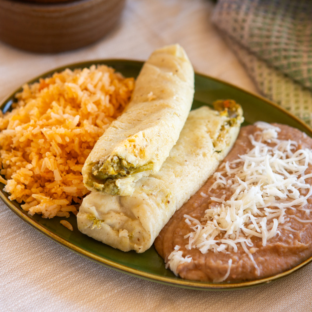 A plate with a cheese-topped burrito next to servings of orange rice and refried beans sprinkled with shredded Hatch Green Chile Veggie Tamales, placed on a beige tablecloth.