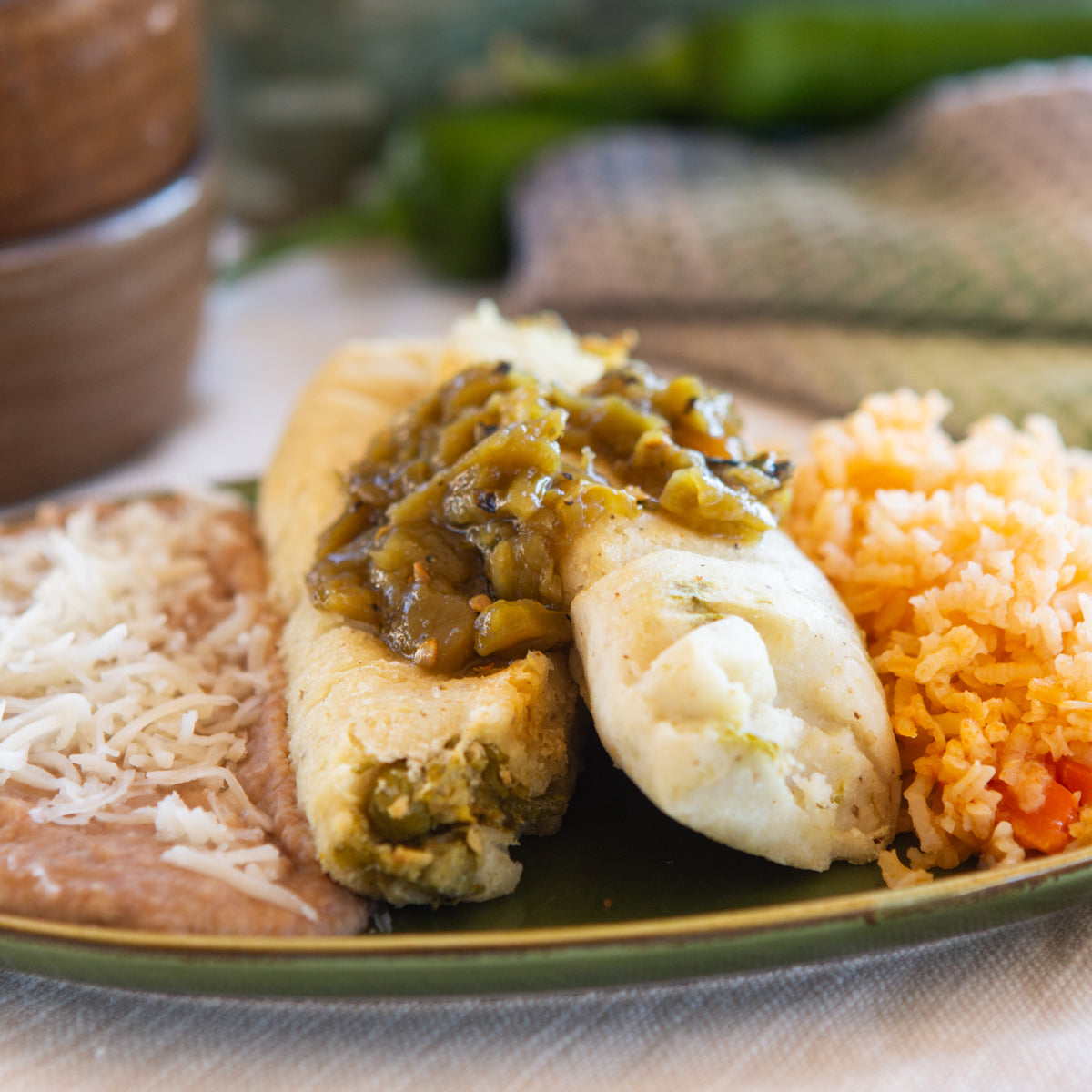 A plate with a Hatch Green Chile Veggie Tamales topped with Hatch Green Chile sauce, accompanied by Spanish rice and refried beans, presented on a textured tablecloth.