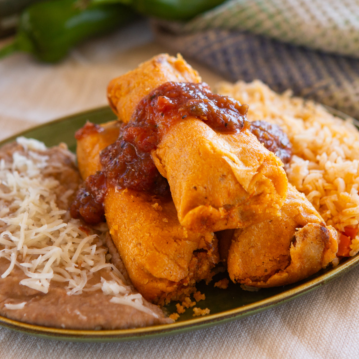 A plate of Mexican food featuring golden-browned Hatch Red Chile Pork Tamales topped with Hatch Red Chile salsa, accompanied by a side of rice and refried beans, garnished with