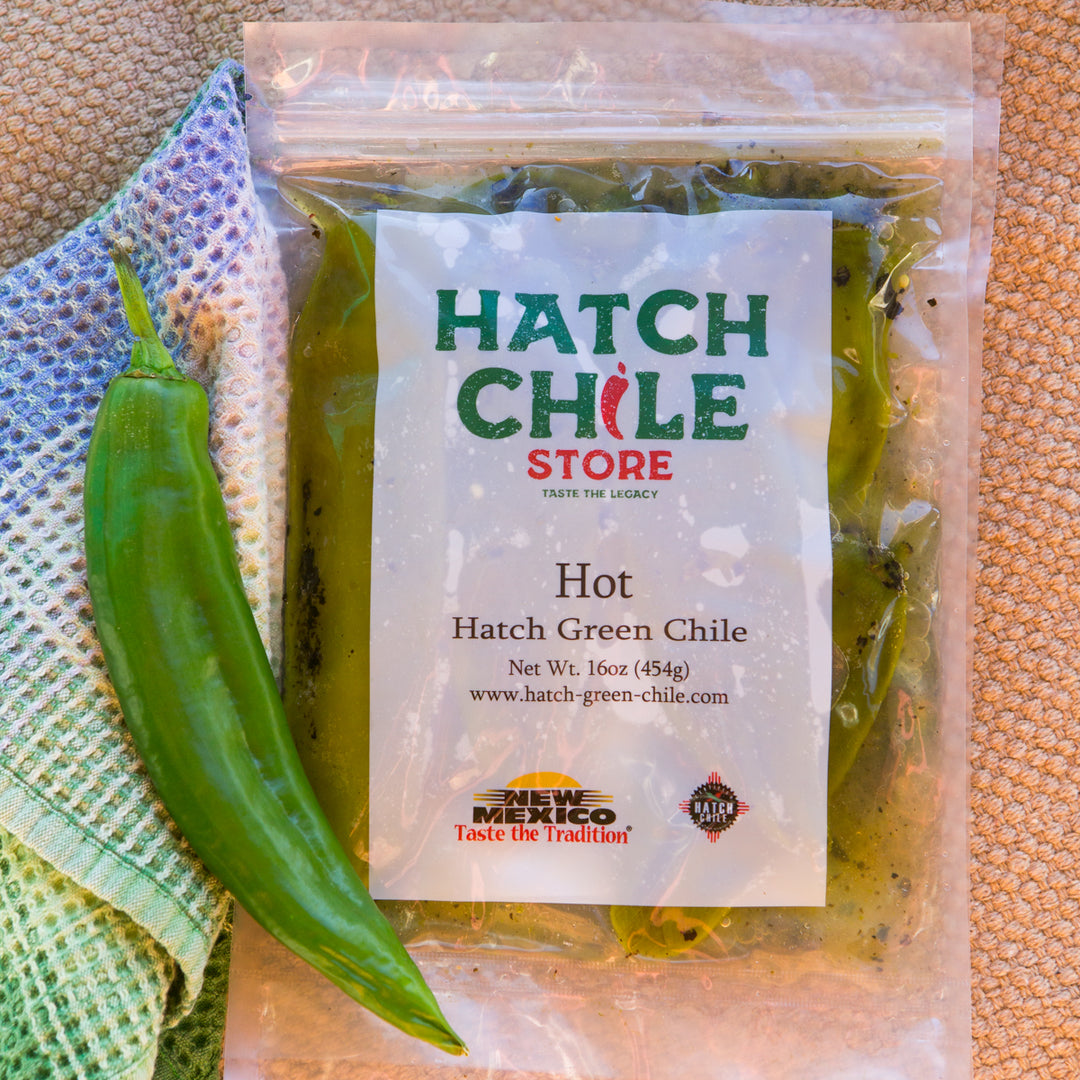 A package of Roasted Hatch Green Chile labeled "hot" alongside a fresh green chile pepper and a colorful woven cloth, placed on a textured beige background.