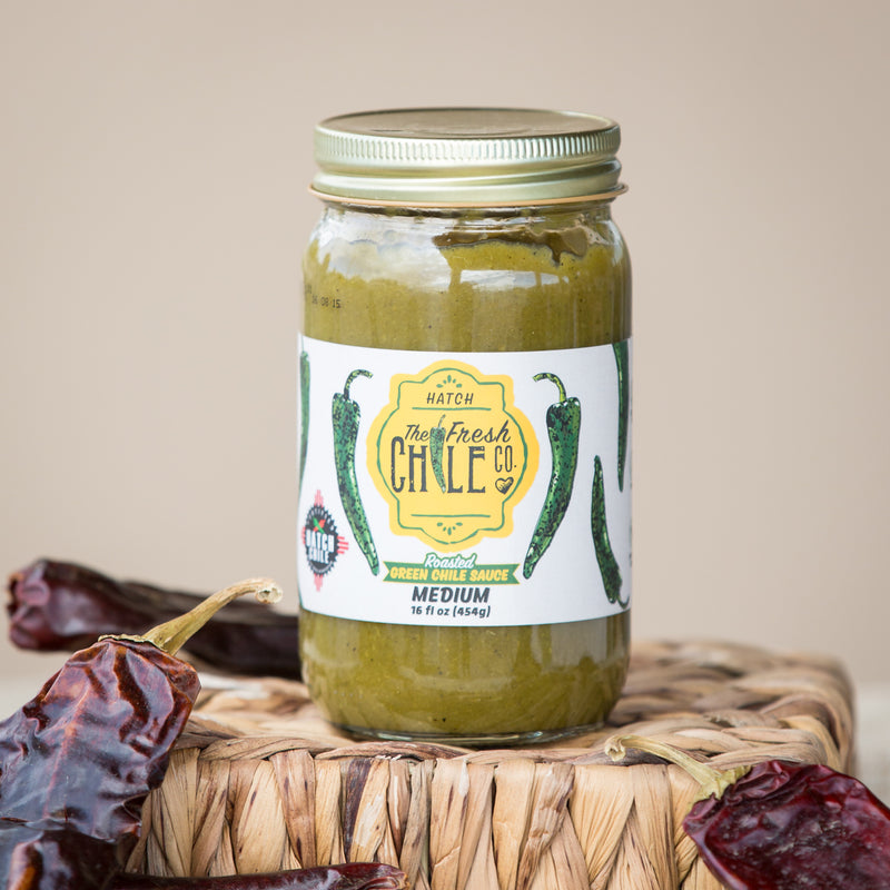 A jar of Roasted Hatch Green Chile Sauce labeled "medium" sits on a woven basket, flanked by dried chili peppers, against a neutral background.