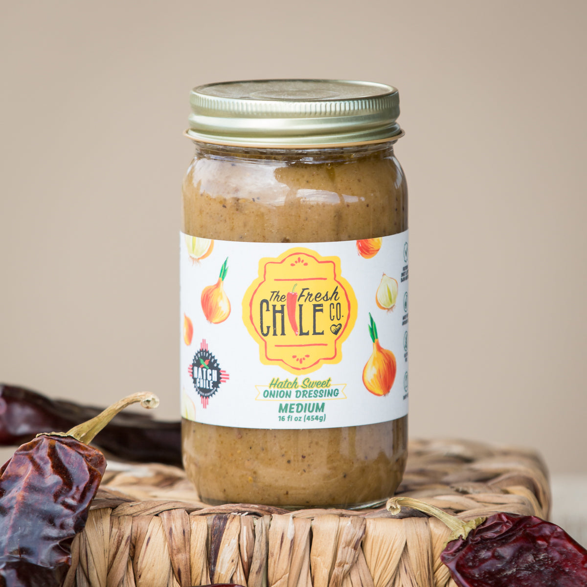 A jar of Hatch Sweet Onion Dressing by The Fresh Chile Co., labeled "medium," placed on a woven basket, surrounded by dried red chili peppers, against a beige backdrop.