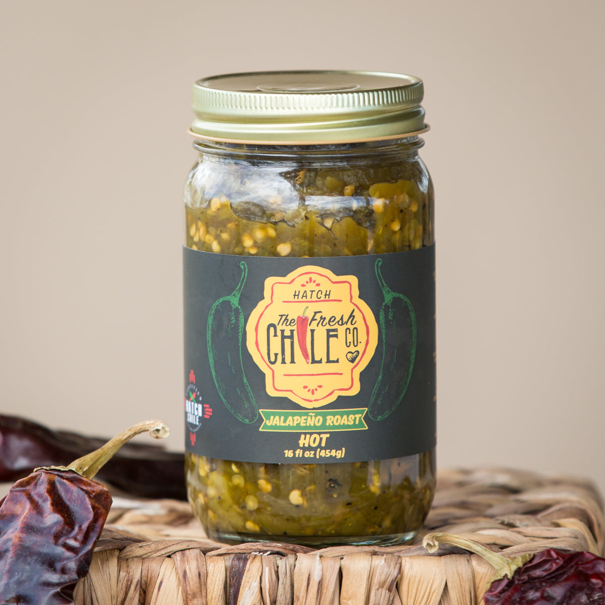 A jar of Hatch Green Jalapeño Roast pickles sits on a wicker surface, flanked by dried red chilies, with a light beige background.