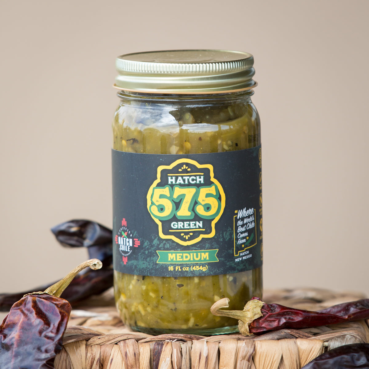 A jar of Hatch 575 Green Chile Sauce labeled "medium" on a table, surrounded by dried chili peppers, with a warm beige background.