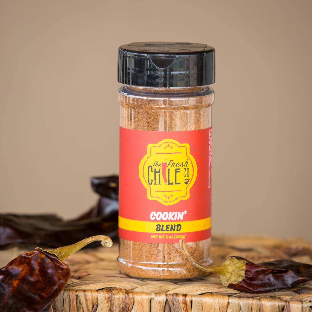 A jar of Cookin' Blend seasoning by "the fresh chile co.", surrounded by dried chile peppers, on a beige background. The label is red with bold yellow text.