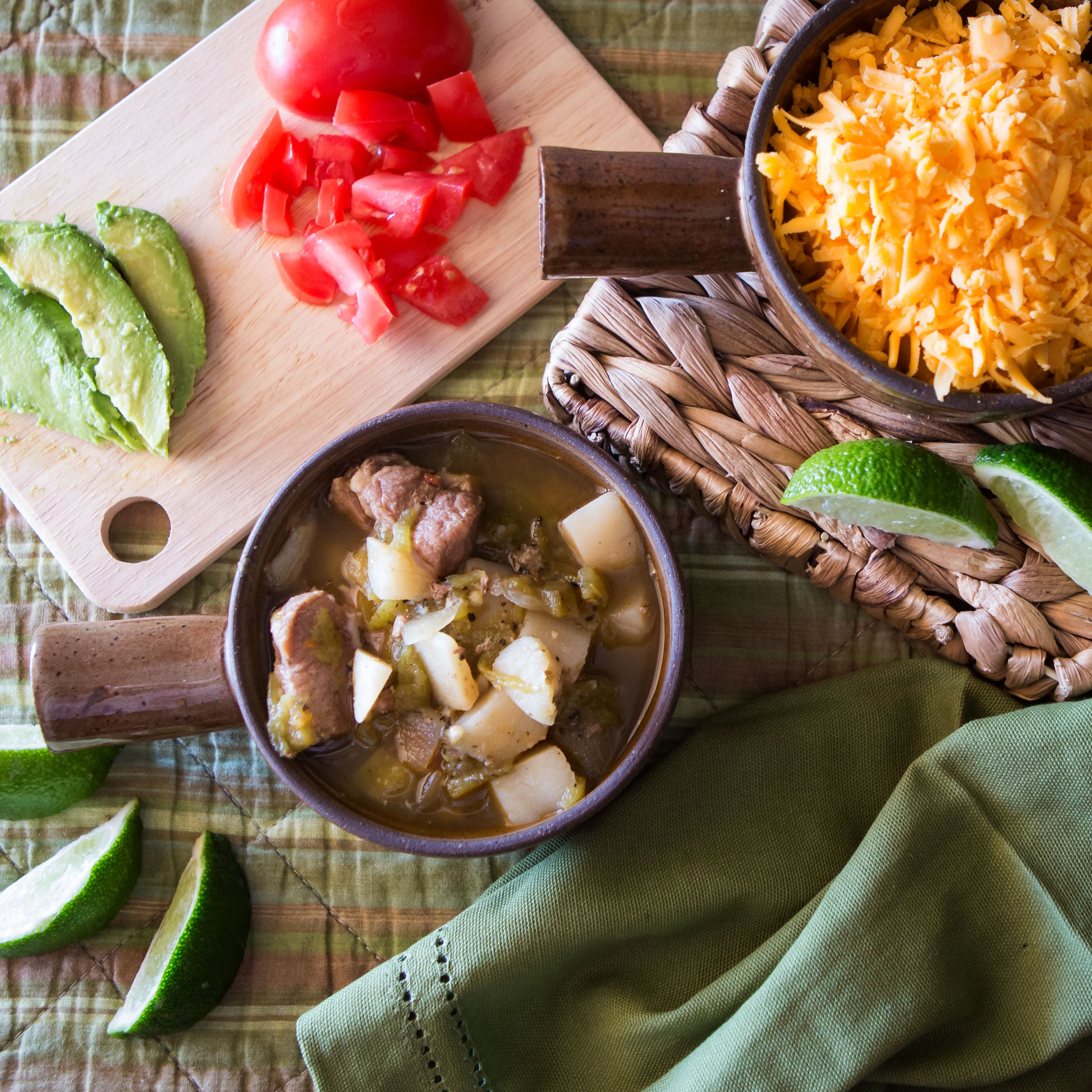 A rustic table setting featuring a bowl of New England Clam Chowder with meat and potatoes, sliced avocado, chopped tomatoes, and shredded cheese, emphasizing a hearty, homemade meal.