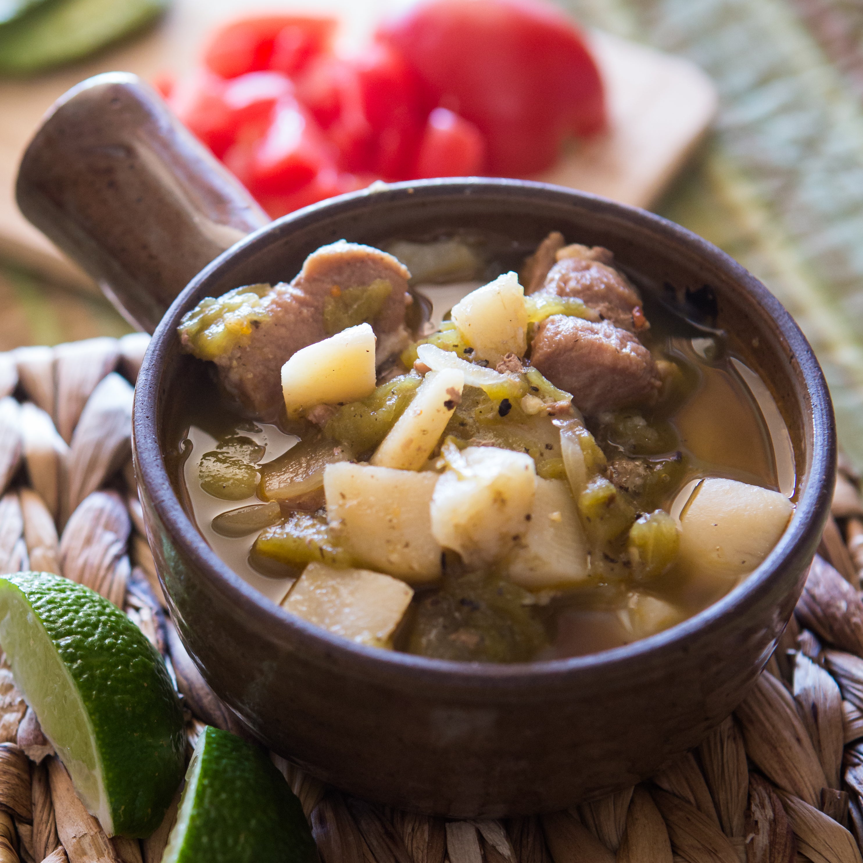 A bowl of traditional Green Chile Stew with chunks of meat and potatoes, garnished with lime wedges, served on a woven mat with tomatoes in the background.