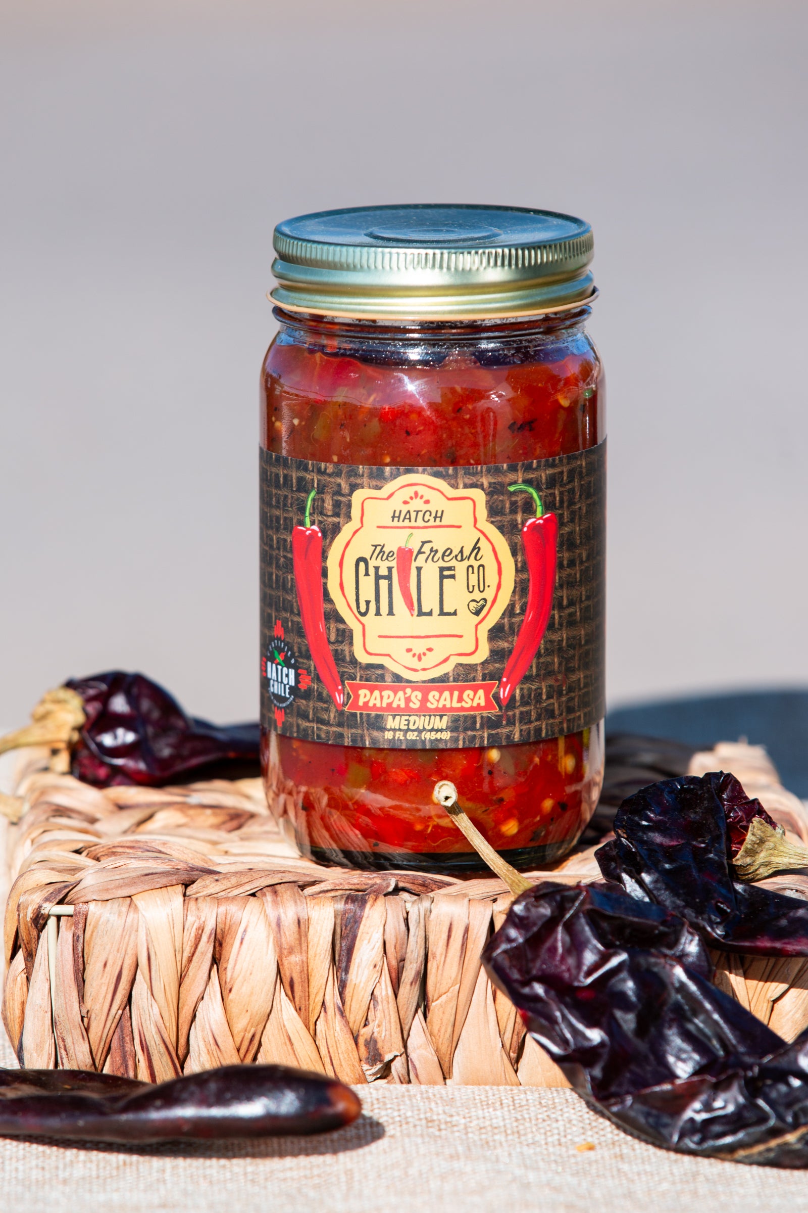 A jar of medium sampler, labeled "papa's salsa - medium," with dried chiles around it on a woven basket, against a neutral backdrop.