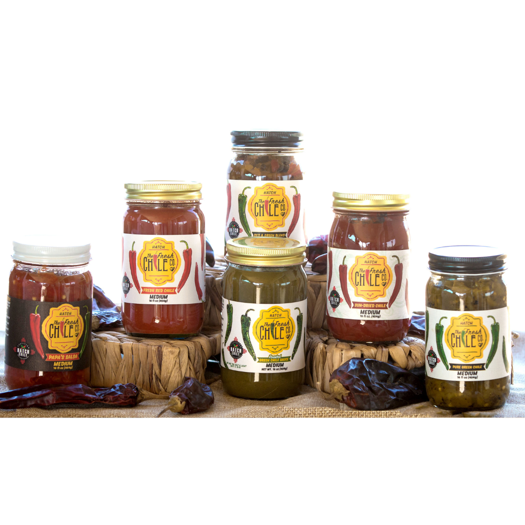 A line of five Medium Sampler jars of chili lee salsa and sauces, including Hatch Red Chile Sauce and Hatch Green Chile Sauce, arranged side by side on a woven mat. Each jar has a label featuring the brand