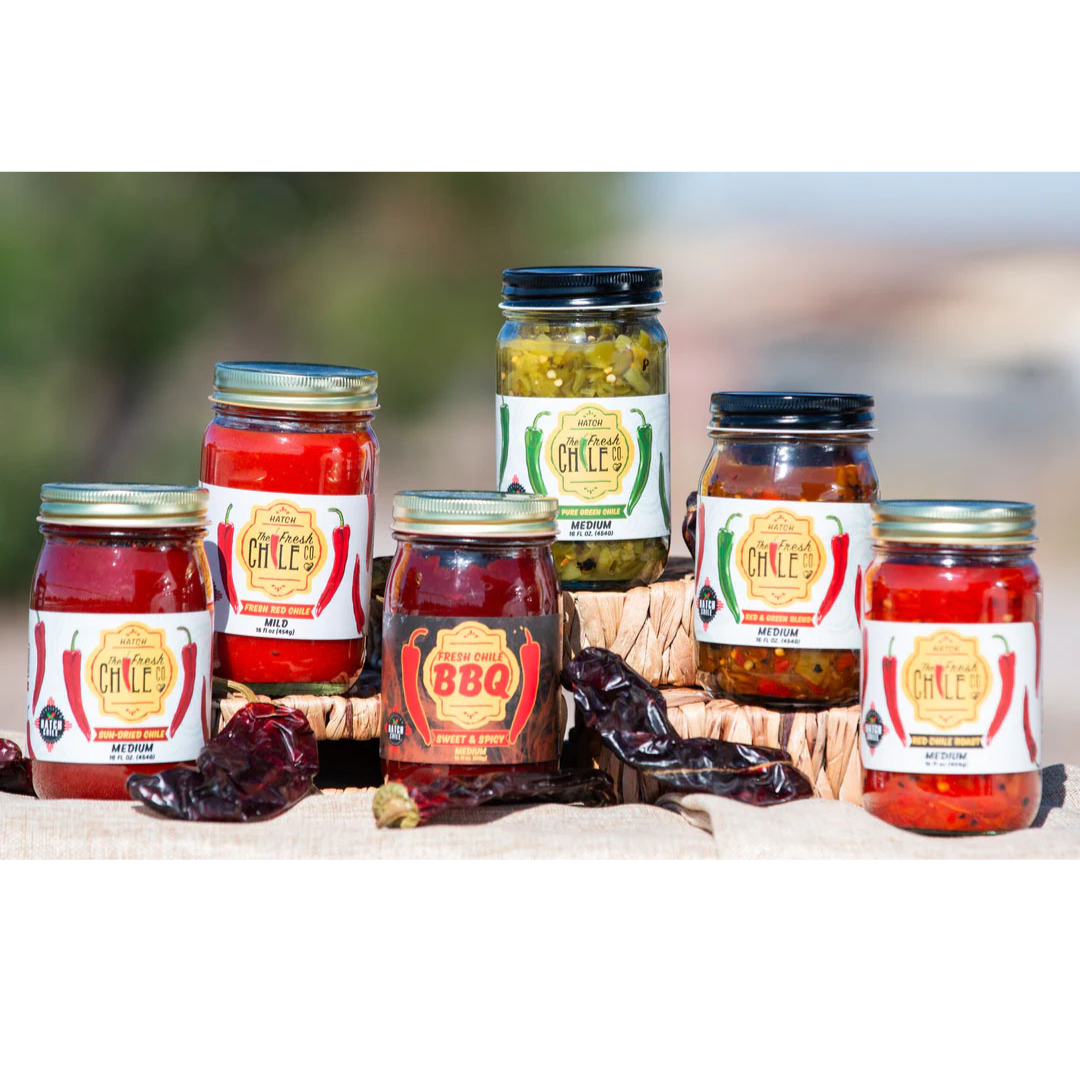 Five jars of Sauces & Roasts Sampler brand products, including Hatch Chile Sauces, displayed outdoors featuring salsas and pickled vegetables with dried chili peppers and a natural backdrop.