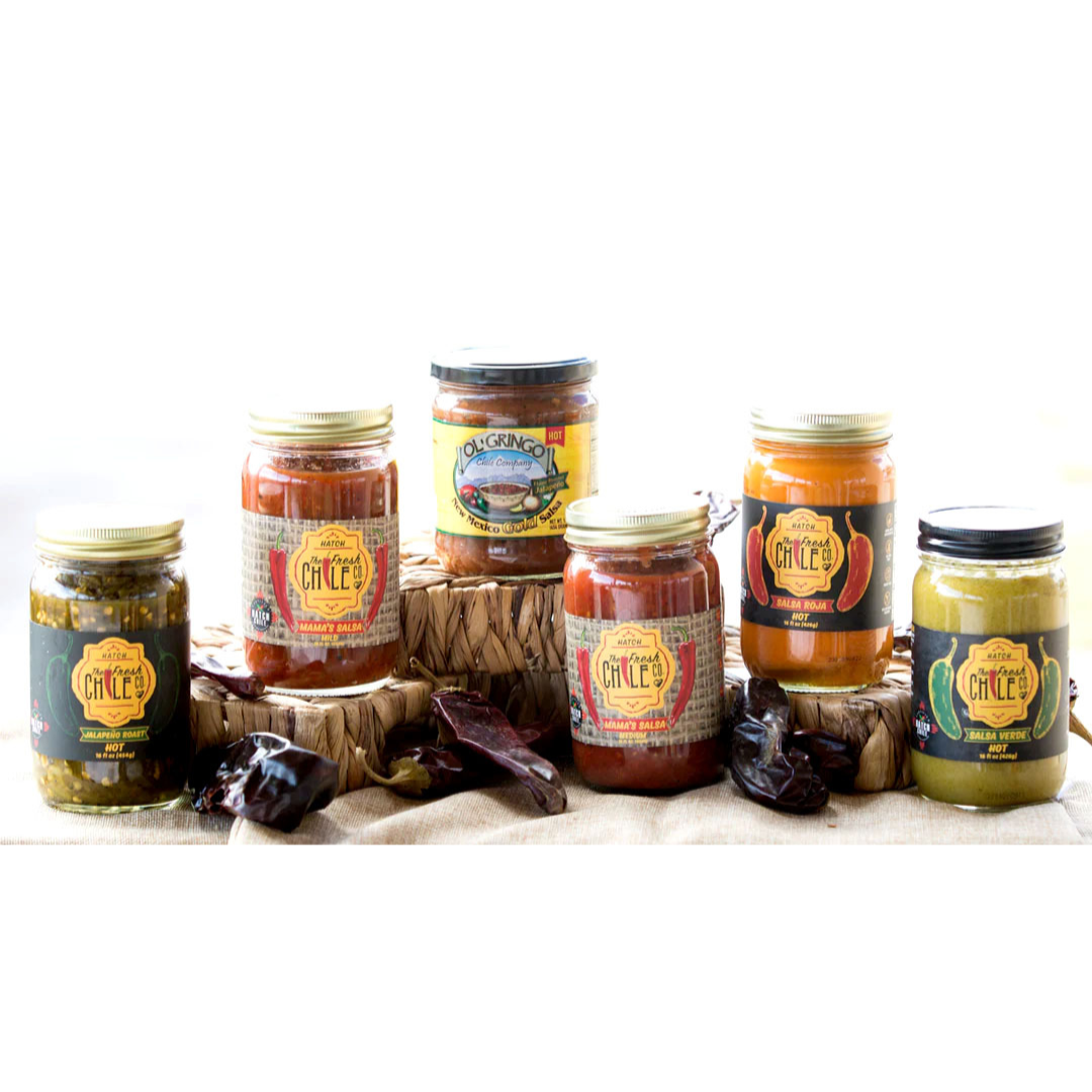 Five jars of Hot Salsa Sampler brand salsas in a variety gift pack on a woven basket against a bright background, including varieties of green and red salsa, flanked by dried chili peppers.