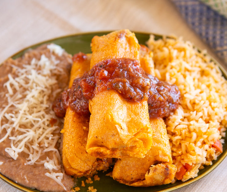 Plate of three Hatch Red Chile Pork Tamales topped with Hatch Red Chile sauce, accompanied by sides of Mexican rice and refried beans, garnished with shredded cheese, on a green plate.