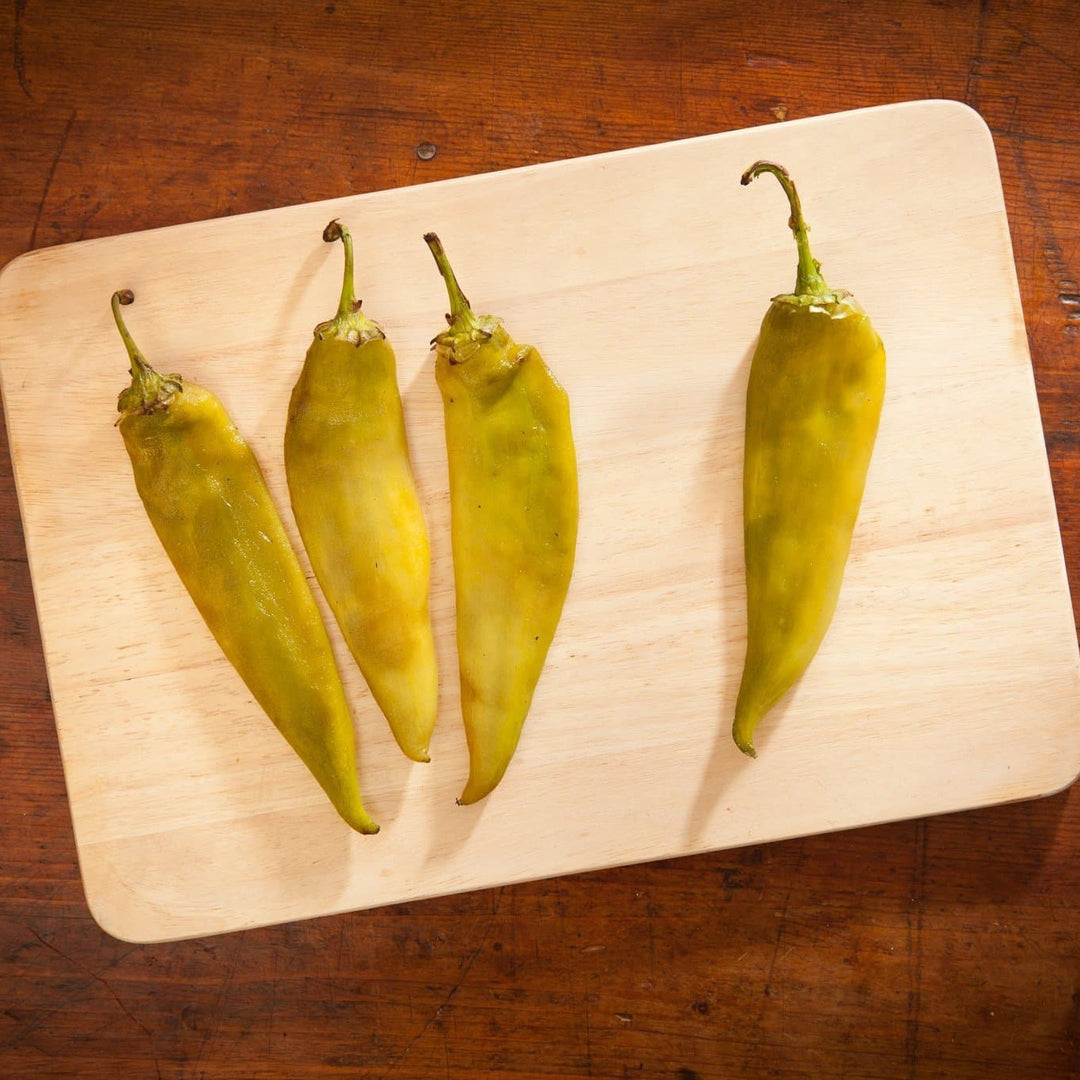 Four Roasted Hatch Green Chile peppers laid out on a wooden cutting board, displaying varying degrees of ripeness and texture.