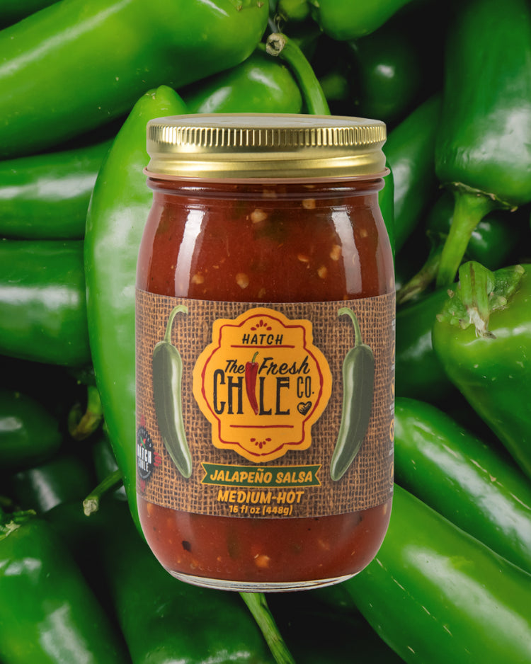 A jar of Hot Salsa Sampler labeled "medium hot," surrounded by fresh green jalapeños, emphasizing its main ingredient from the variety of New Mexican salsas.