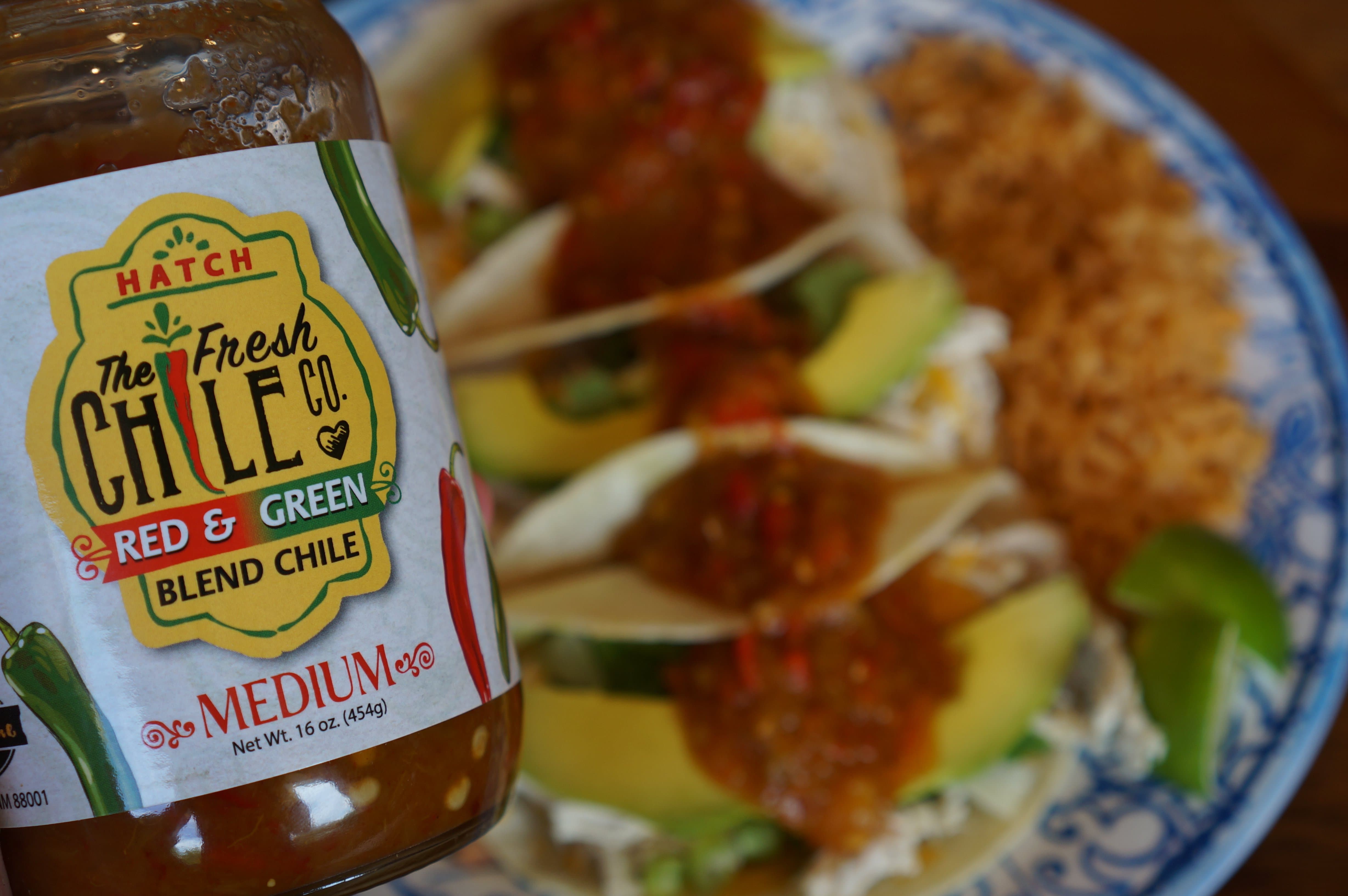 Sentence with replaced product: Close-up of a meal with fish tacos garnished with slices of avocado and a medium 505 Southwestern Hatch Red & Green Chile sauce, served on a decorative blue plate with a side of brown rice.