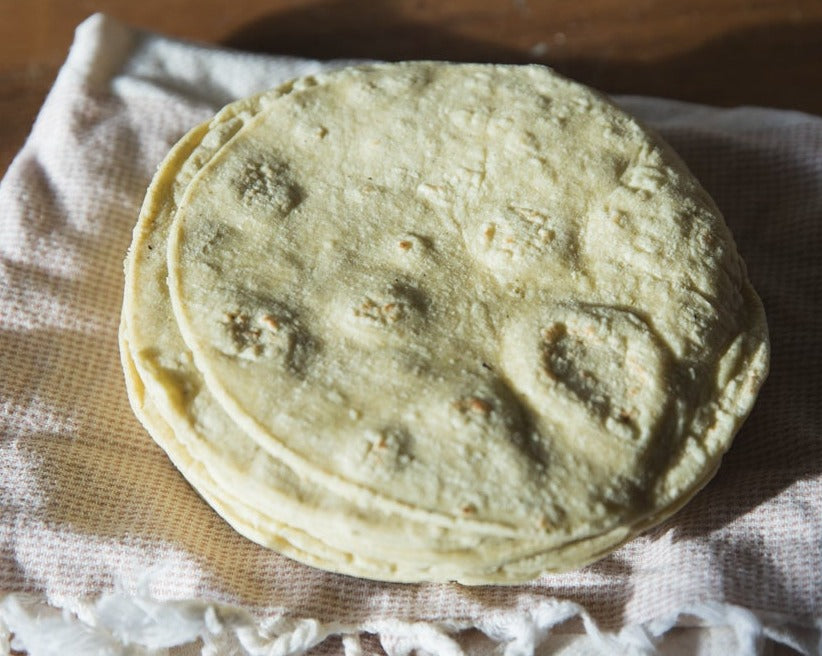 A stack of freshly made Hatch Green Chile corn tortillas on a white cloth with a wooden surface underneath. The Green Chile Corn Tortillas have a slightly golden texture with visible char spots.