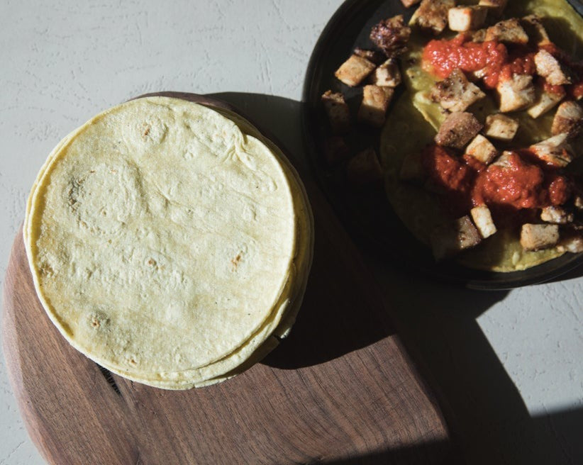 A stack of Green Chile Corn Tortillas on a wooden board next to a bowl of chilaquiles topped with chunky salsa, placed on a textured white surface under sunlight.