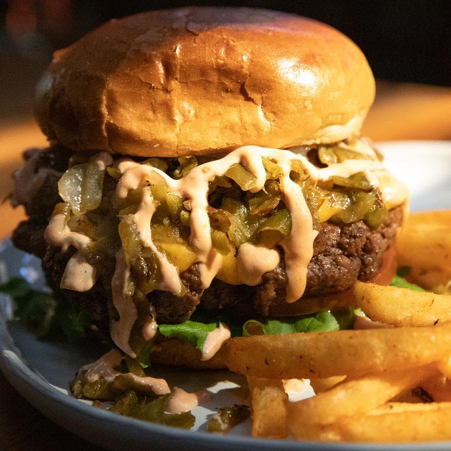 A juicy hamburger with lettuce, pickles, Pure Hatch Green Chile, and sauce on a shiny bun, served with a side of golden fries on a white plate, highlighted under warm lighting.