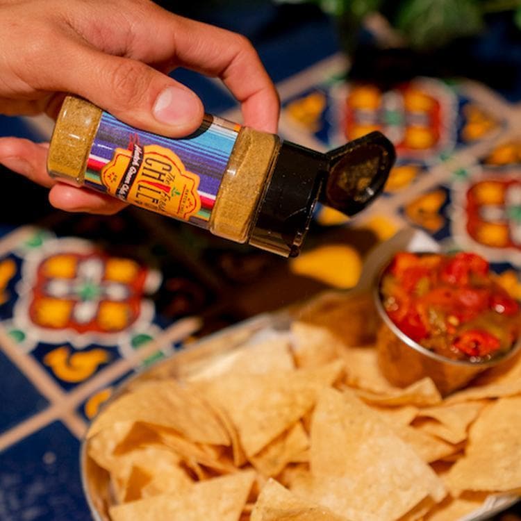 A person's hand pouring salsa seasoned with Hatch Green Chile Spice from a colorful jar onto a bowl of nachos, with fresh ingredients and a dish of diced tomatoes in the background.