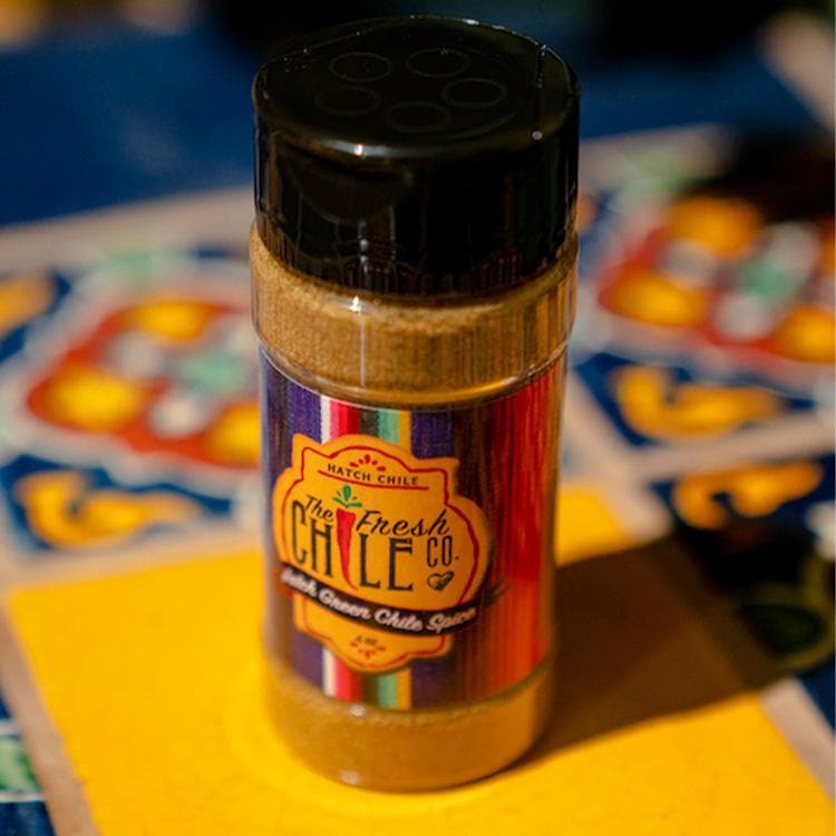 A container of Hatch Green Chile Spice from the fresh chile co., resting on a yellow napkin with a colorful patterned tablecloth in the background.