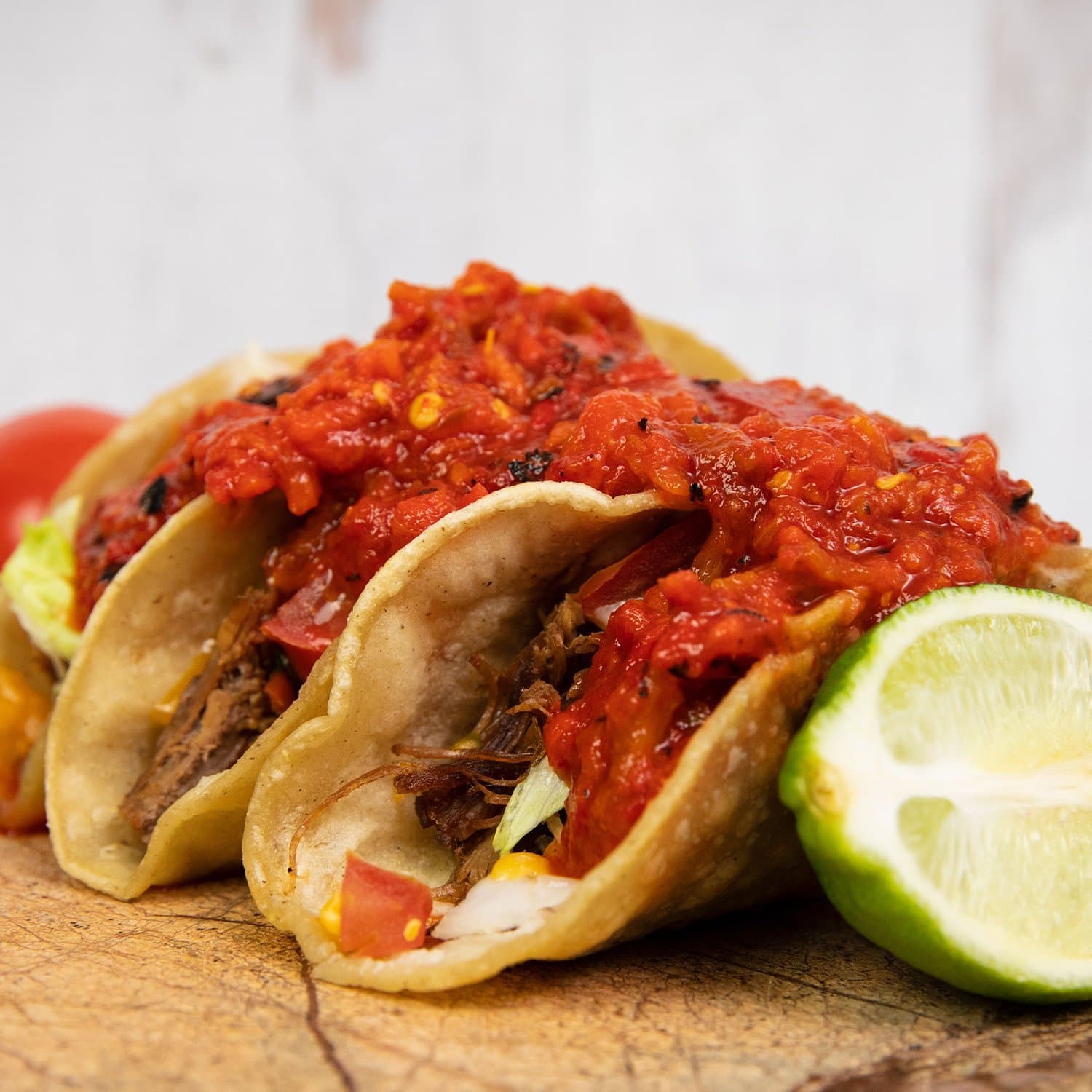 Three gluten-free tacos filled with Hatch Red Chile Roast and topped with salsa on a wooden surface, garnished with lime wedges and a blurred tomato in the background.