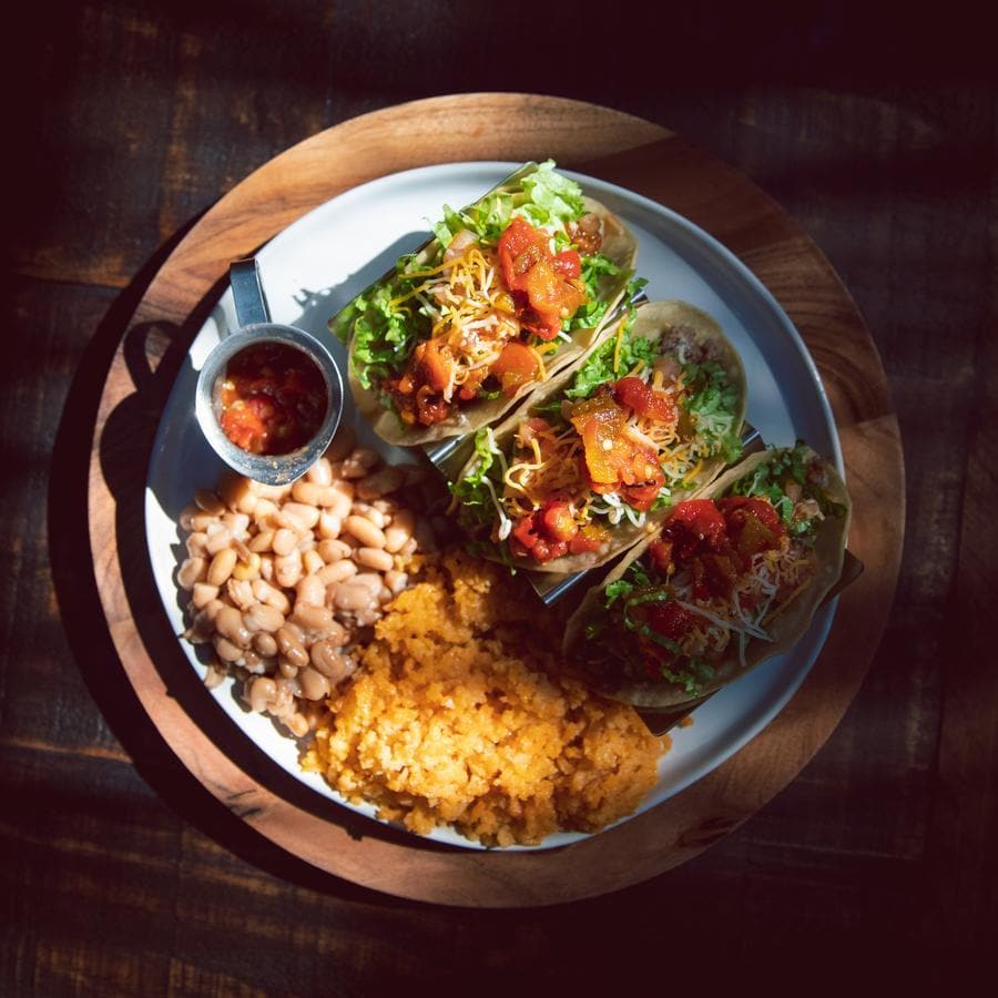 A plate with tacos, rice, beans, and Fresh Chile Co's Mama's Blended Hatch Chile Salsa served on a wooden tray under sunlight, creating stark shadows and highlights on the food.