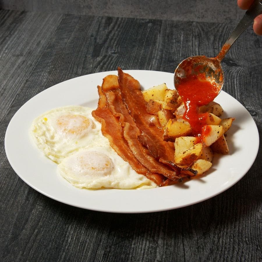 A breakfast plate with two sunny-side-up eggs, crispy bacon strips, and diced potatoes, with a hand pouring Fresh Hatch Red Chile Sauce over the meal, on a dark wooden table.