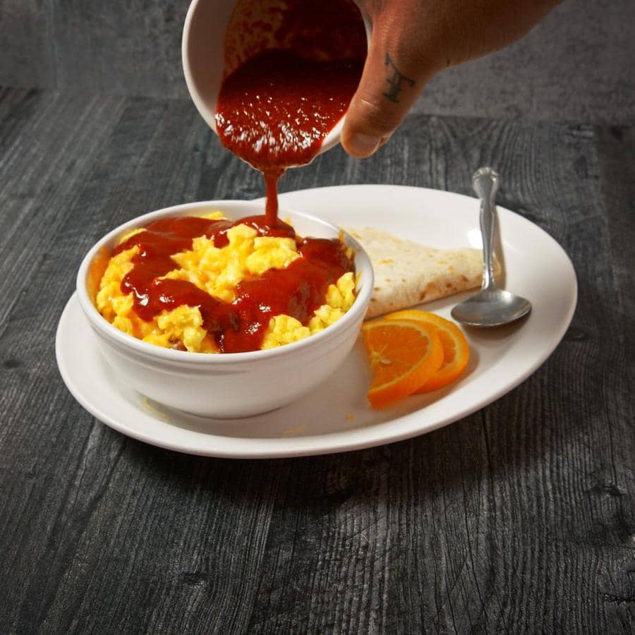 A person pours Sun-Dried Red Chile Sauce over scrambled eggs in a bowl beside a tortilla and orange slices on a dark wooden table.