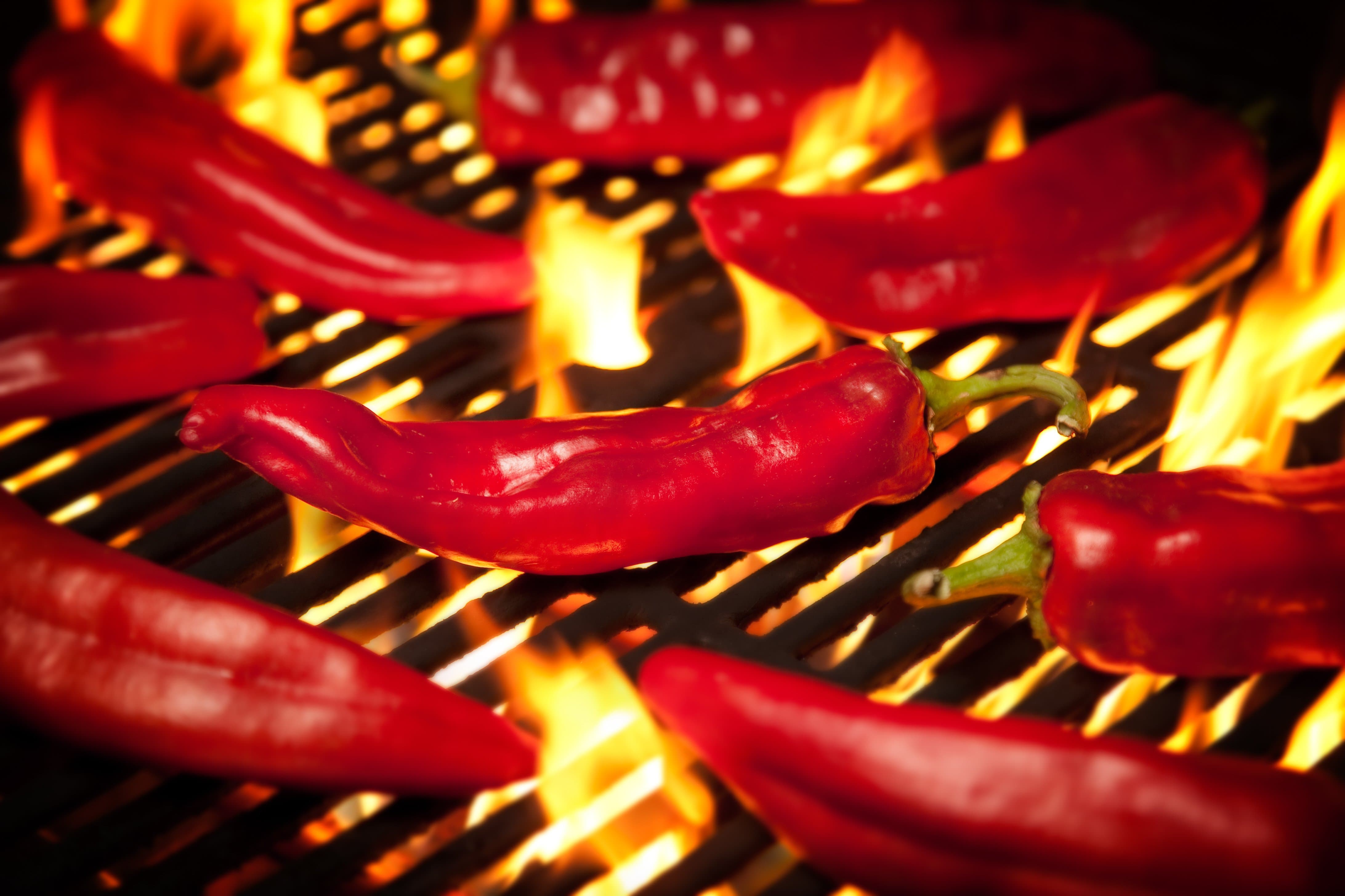 Red chili peppers grilling on a barbecue, with vibrant flames licking the underside of the grill, highlighting the glossy texture of the Fresh Hatch Red Chile Sauce-coated peppers.