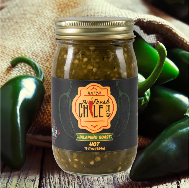A jar of Hatch Green Jalapeño Roast labeled "hot", surrounded by fresh jalapeño peppers on a rustic wooden surface.