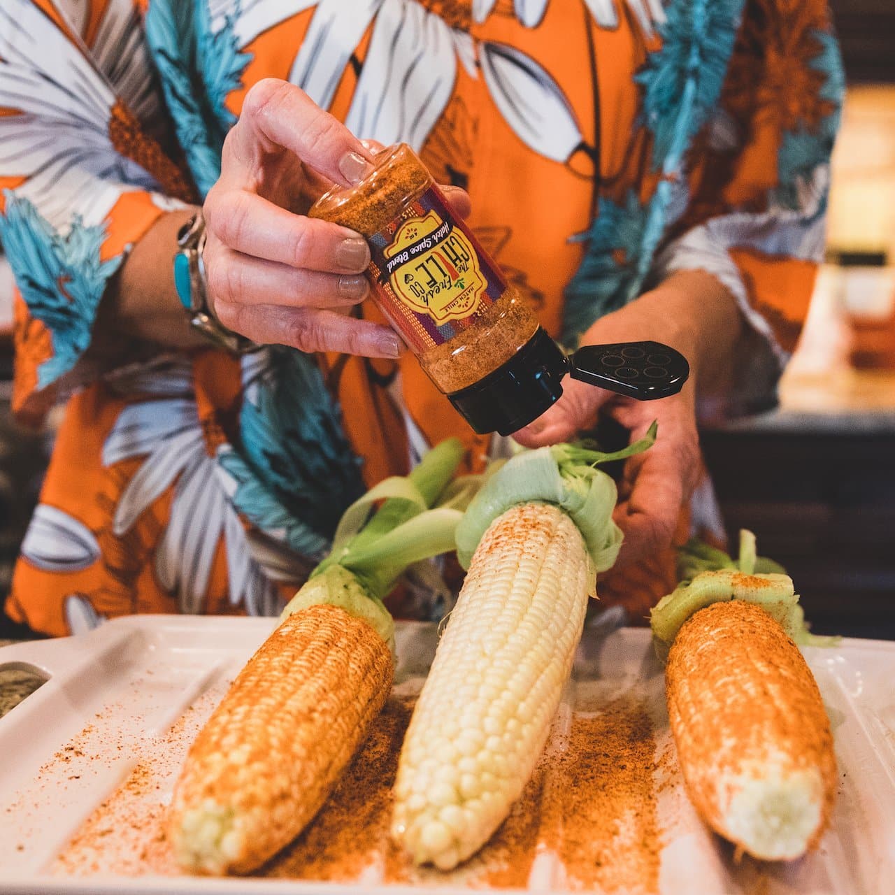 A person in a colorful shirt sprinkles Hatch Red Chile Spice Blend from a bottle onto fresh corn cobs laid out on a tray.