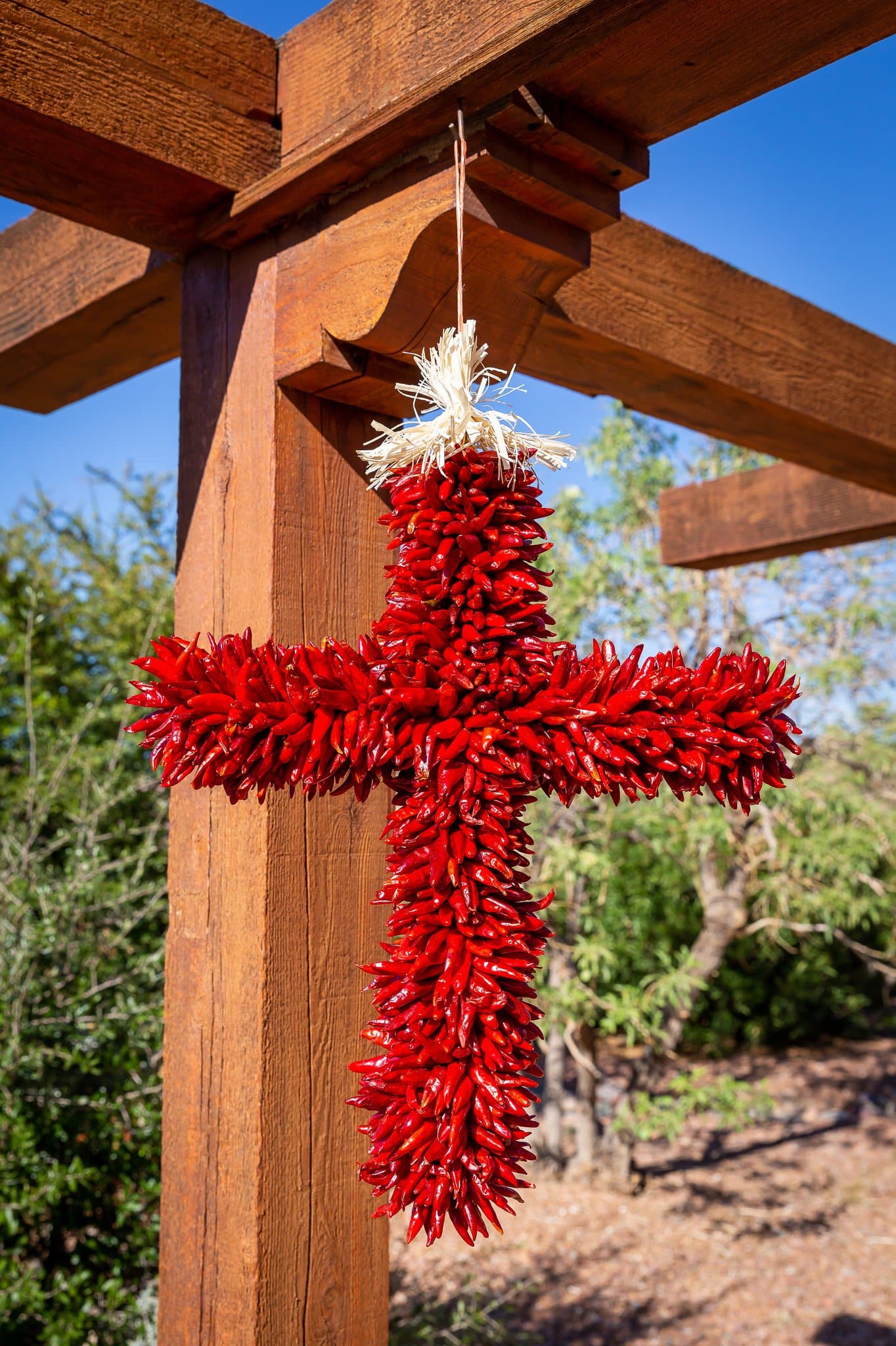 A vibrant red Chile Pequin Cross, hand-made and hanging from a wooden beam under a clear blue sky, symbolizing southwestern cultural heritage.