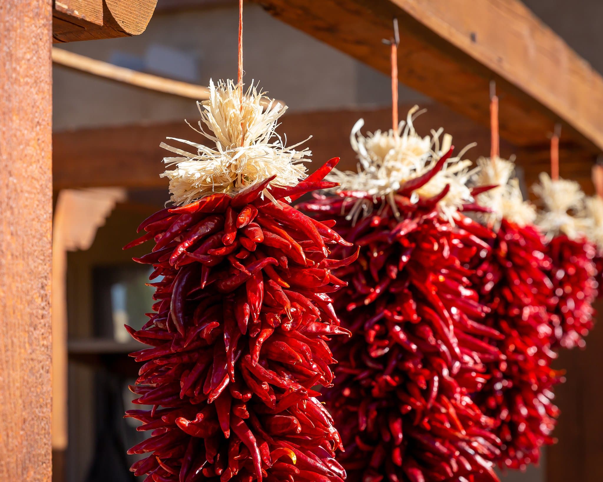 Chile Pequin ristras of red chili peppers hanging from a wooden beam, with straw tied at the top of each bunch, set against a soft-focused building background.