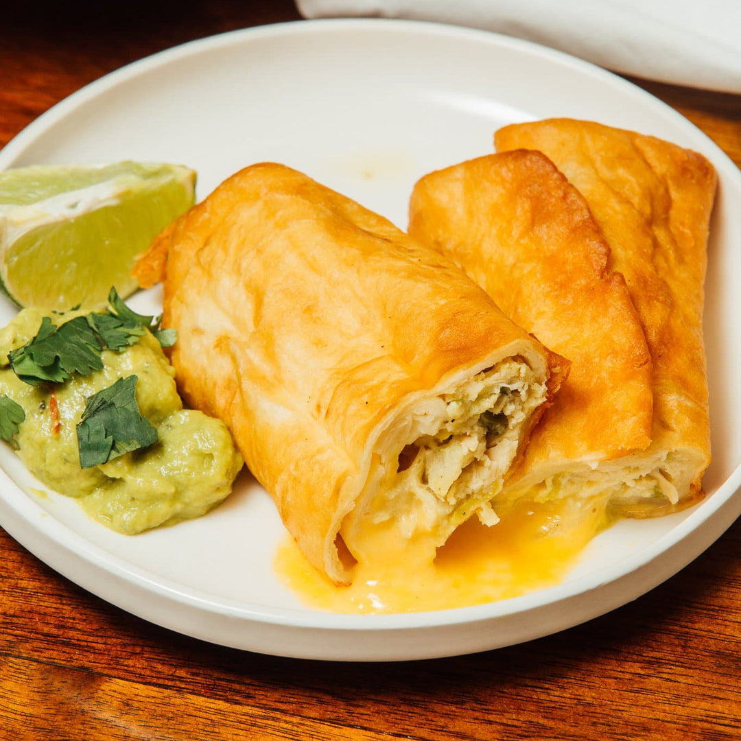 Two crispy golden Hatch Green Chile Beef & Cheese Chimichangas on a white plate, one opened to reveal a Hatch Green Chile Beef & Cheese filling, served with a side of guacamole and a wedge of lime.