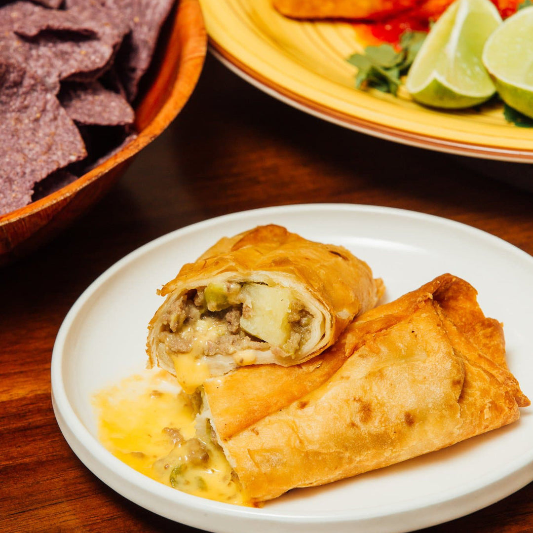 Close-up of Hatch Green Chile Beef & Cheese Chimichangas on a plate with cheese sauce, accompanied by a bowl of blue corn tortilla chips and lime wedges, on a wooden table.