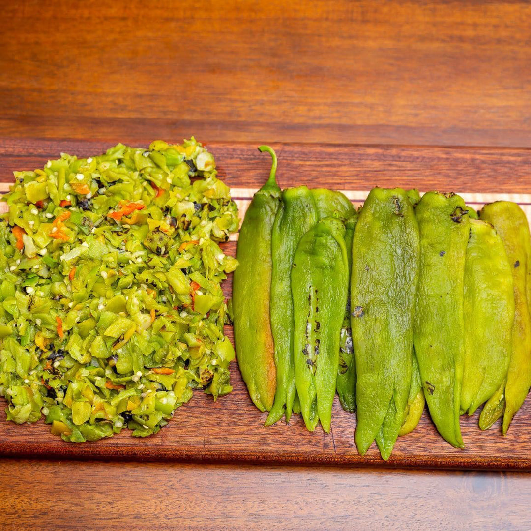 Sentence with product name: A wooden platter holds chopped mixed vegetables on the left and Roasted Hatch Green Chile peppers on the right, all on a dark wooden table background.