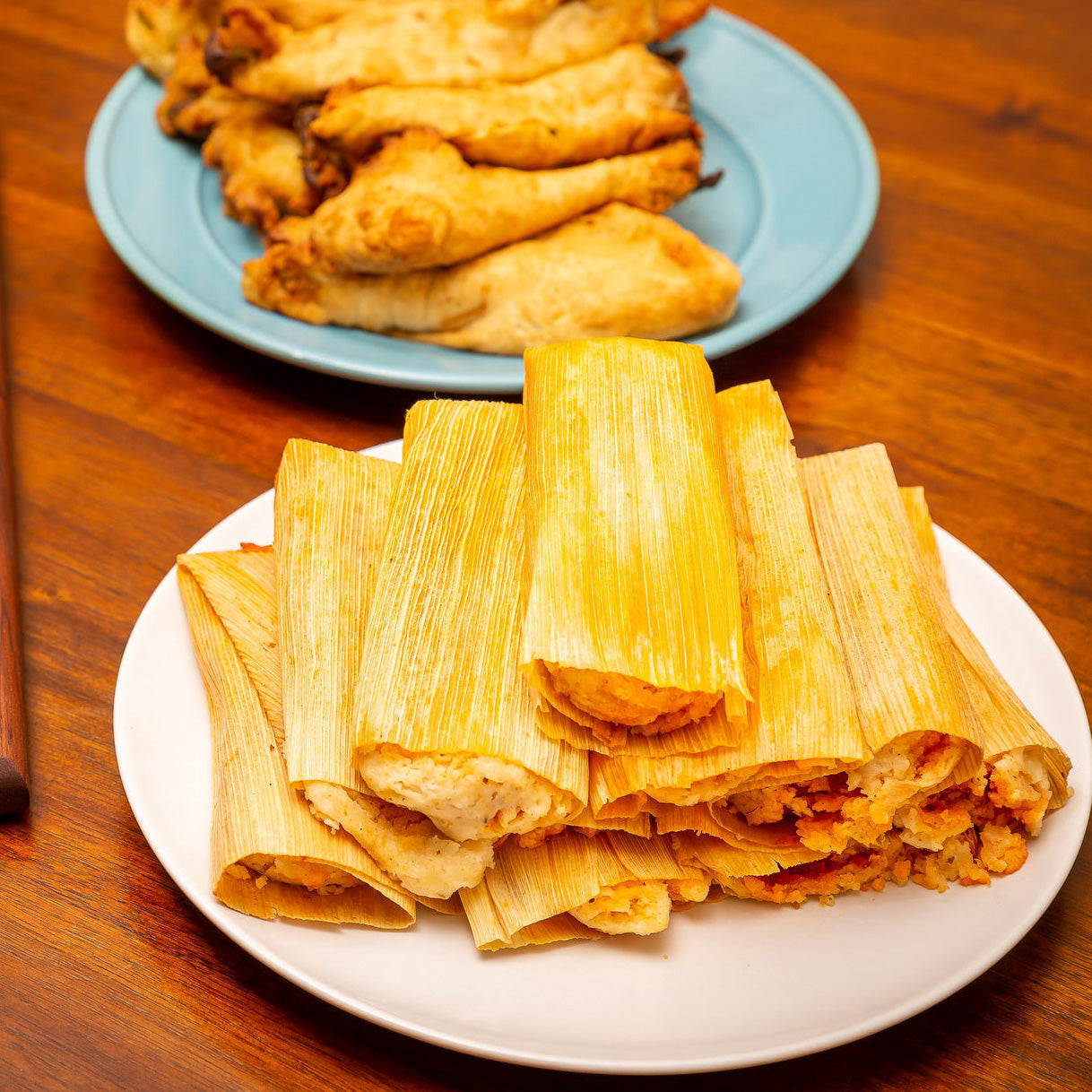A plate of golden-brown Hatch Green Chile Veggie Tamales neatly stacked, served alongside a dish of empanadas on a wooden table.