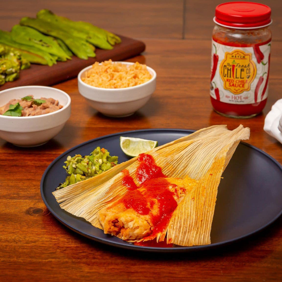 A plate with Hatch Red Chile Pork Tamales topped with Hatch Red Chile sauce, accompanied by lime wedges, served with sides of rice, beans, and nopales in the background, next to a jar