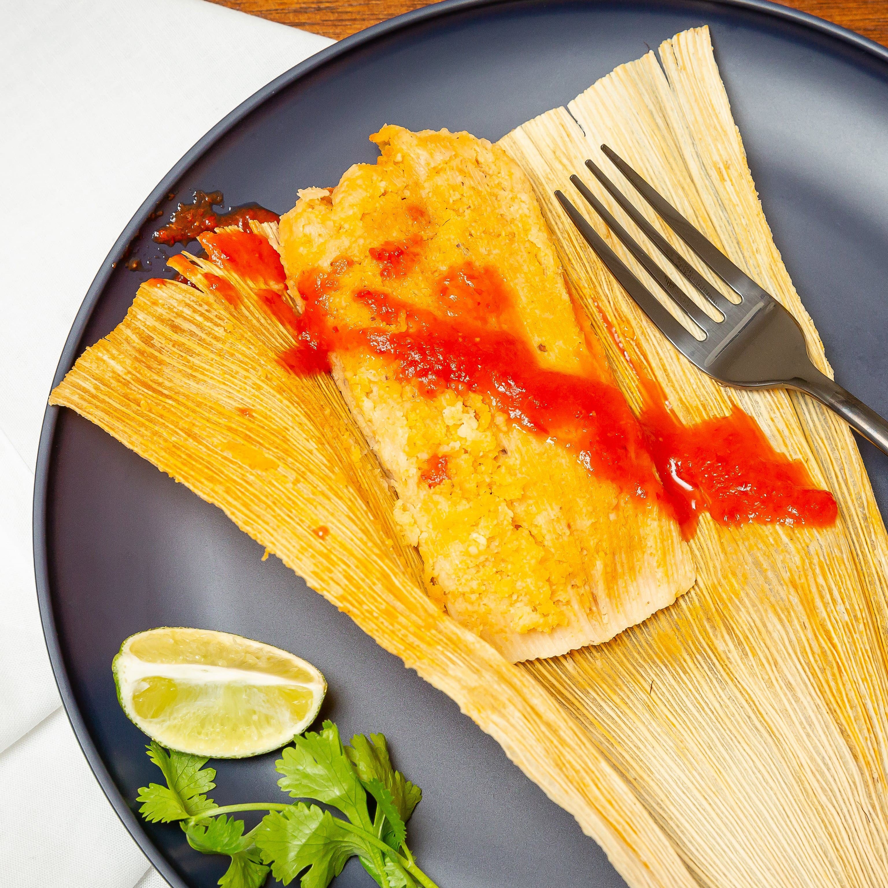 A freshly cooked Hatch Green Chile Chicken Tamale with bright red sauce on top, placed on a dark blue plate accompanied by a slice of lime and a sprig of cilantro, with a fork resting.